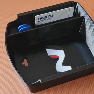 Könnte beinhalten: Schwarzer Kunststoff-Autokonsolen-Organizer mit Fächern. Enthält einen Schlitz für eine Karte mit der Aufschrift "NESTE", ein kleines blaues Objekt und eine silberne und rote Grafik. Der Organizer befindet sich auf einer braunen Oberfläche.
