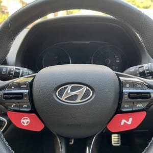 Può includere: Un volante in pelle nera con il logo Hyundai argentato al centro. Accenti rossi sui lati sinistro e destro del volante. Lo sfondo mostra il cruscotto dell'auto e uno scorcio dell'esterno.