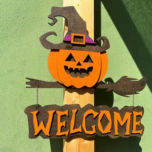 Puede incluir: Un letrero de bienvenida de Halloween de madera con una calabaza que lleva un sombrero de bruja y sostiene una escoba. El letrero dice "WELCOME" en letras naranjas.