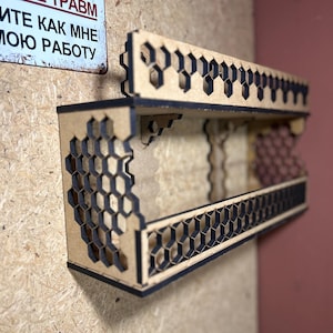 Puede incluir: Una estantería de madera con un diseño de panal. La estantería está hecha de madera marrón claro con detalles negros. La estantería está montada en una pared.