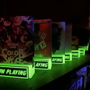 May include: A row of illuminated book displays with the words "NOW PLAYING" in green. The book covers feature titles like "COLOR OUT OF SPACE" and "BURN OK". The displays are on a shelf.