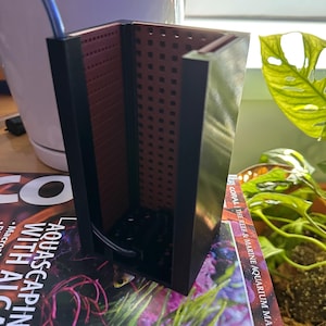 Könnte beinhalten: Ein rechteckiger, schwarzer und roter Aquariumfilter mit perforiertem Innenraum. Der Filter befindet sich auf einem Magazin mit dem Titel "Aquascaping with Algae". Ein blauer Schlauch erstreckt sich vom Filter.