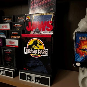 May include: A display stand featuring vintage video game boxes. The boxes include "Star Voyager," "Jaws," and "Jurassic Park." The stand has a controller design at the base. Other Atari game cartridges are visible in the background.