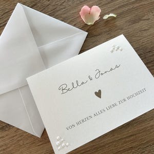 Könnte beinhalten: Eine weiße Hochzeitseinladung mit den Namen "Bella & Jonas" und einem Herzsymbol. Der Text "VON HERZEN ALLES LIEBE ZUR HOCHZEIT" ist darunter gedruckt. Ein weißer Umschlag und ein rosa Rosenblatt sind ebenfalls vorhanden.