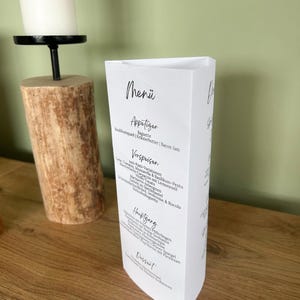 May include: A white, triangular menu card with the word "Menü" at the top, followed by "Appetizer" and a list of food items. The menu is on a wooden surface, with a candle and wooden candle holder in the background.