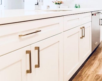 May include: White kitchen cabinets with brushed gold hardware. The cabinets have a simple, modern design and are installed in a row.