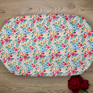 Puede incluir: Una sábana bajera blanca de forma ovalada para cuna con un estampado floral de flores rojas, amarillas y azules sobre fondo blanco.