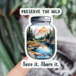 May include: A sticker featuring a jar with a landscape scene of mountains, trees, and a lake. The text "PRESERVE THE WILD" is above the jar, and "Save it. Share it." is below. The sticker is held by a hand.