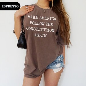 May include: A brown oversized t-shirt with the text "Make America Follow the Constitution Again" printed in white. The shirt is being worn by a person with long brown hair and denim shorts.