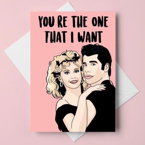 Può includere: Biglietto di auguri rosa con la scritta "YOU RE THE ONE THAT I WANT". Il biglietto presenta un'illustrazione di un uomo e una donna che si abbracciano. La donna ha i capelli biondi e l'uomo ha i capelli neri. Il biglietto è in una busta.