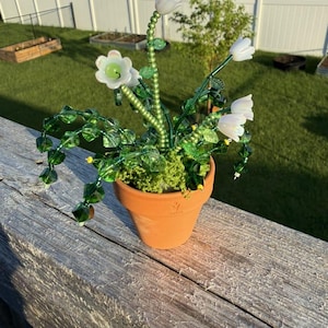 May include: A decorative plant sculpture featuring white glass flower blossoms and green beaded stems and leaves, arranged in a small terracotta pot. The stems are constructed from strings of green beads, and the foliage consists of faceted green glass beads.