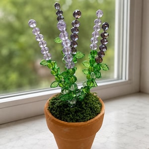 May include: A decorative plant sculpture featuring four stems of faceted purple and lavender crystal beads arranged with green crystal leaves. The stems are anchored in a small, round, terracotta-colored clay pot filled with green moss.