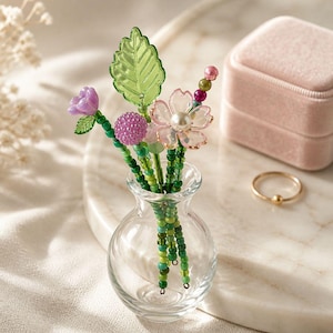 May include: A miniature clear glass vase containing a bouquet of beaded stems. The stems feature green glass leaves, a purple flower, a textured purple sphere, and a light pink flower with a white pearl center, accented by small green beads.