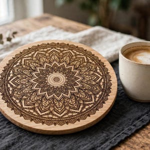 May include: A circular wooden coaster featuring a dark brown laser-engraved mandala pattern with intricate geometric details, symmetrical floral motifs, and a central starburst design, displayed on a dark fabric runner next to a ceramic mug.