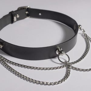 Op de afbeelding: Zwarte leren choker ketting met zilverkleurige kettingaccenten. De choker heeft een gespsluiting en drie hangende kettingen die met metalen ringen zijn bevestigd. Het accessoire is ontworpen voor de nek en wordt op een witte achtergrond getoond.