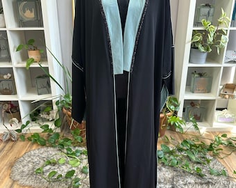 Puede incluir: Una abaya negra con forro azul claro y ribete de cuentas. La abaya tiene cuello en V y mangas largas.