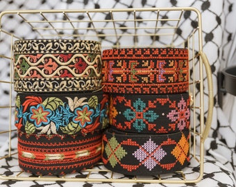 May include: A set of five colorful embroidered trim bands in a gold wire basket. The trim bands are black with various colorful floral and geometric patterns.