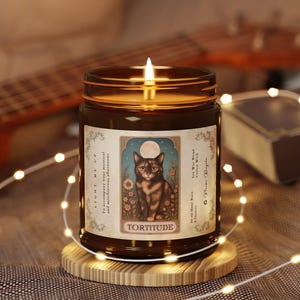 Pode incluir: Um frasco de vela de vidro castanho aceso com um rótulo com uma ilustração de gato, a palavra "TORTITUDE" e o texto "LIGHT ME UP". A vela está sobre um suporte de madeira, com luzes de corda e uma guitarra ao fundo.