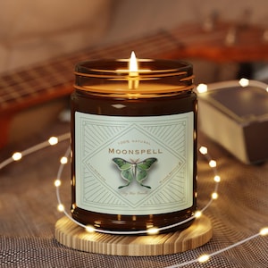 May include: A lit, brown glass jar candle with a label that reads "MOONSPELL" and features a green butterfly illustration. The candle sits on a round wooden coaster, with a string of fairy lights and a guitar in the background.
