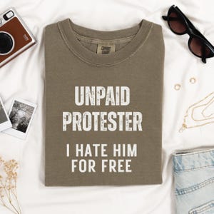 Folded olive green Comfort Colors shirt with protest text, surrounded by Polaroids and accessories.