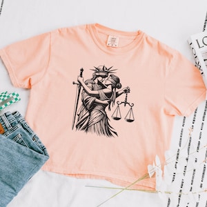 Peachy-pink cropped t-shirt with bold line art of feminist liberty and justice kissing, styled with jeans and graphic book