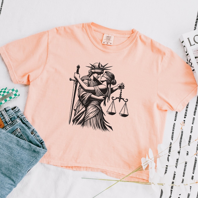 Peachy-pink cropped t-shirt with bold line art of feminist liberty and justice kissing, styled with jeans and graphic book