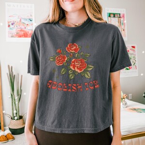 Charcoal cropped t-shirt worn by woman in cozy room with Matisse prints, showing full rose design and groovy text