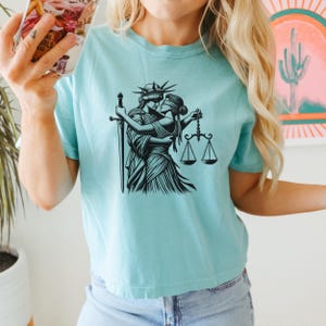 Aqua blue cropped shirt with romantic LGBTQ+ line art of Liberty and Justice about to kiss, modeled while holding phone