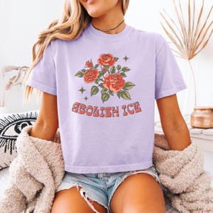 Lavender cropped t-shirt with red roses and bold anti-ICE message, model sitting on bed with cardigan