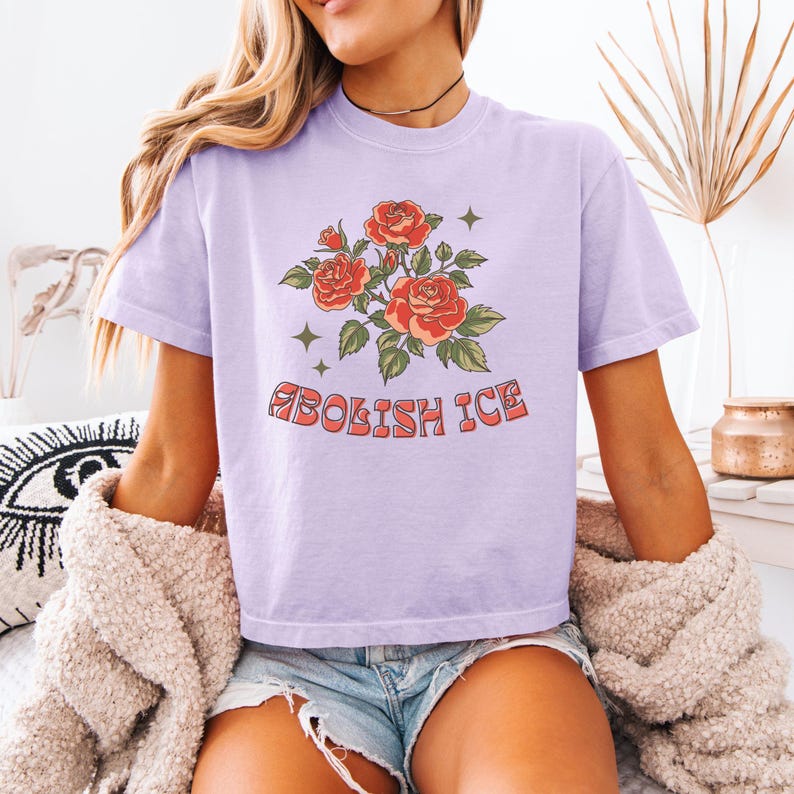 Lavender cropped t-shirt with red roses and bold anti-ICE message, model sitting on bed with cardigan