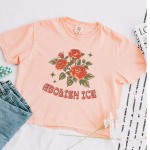 Peach cropped tee with red rose bouquet and groovy abolish ICE text, laid flat with accessories
