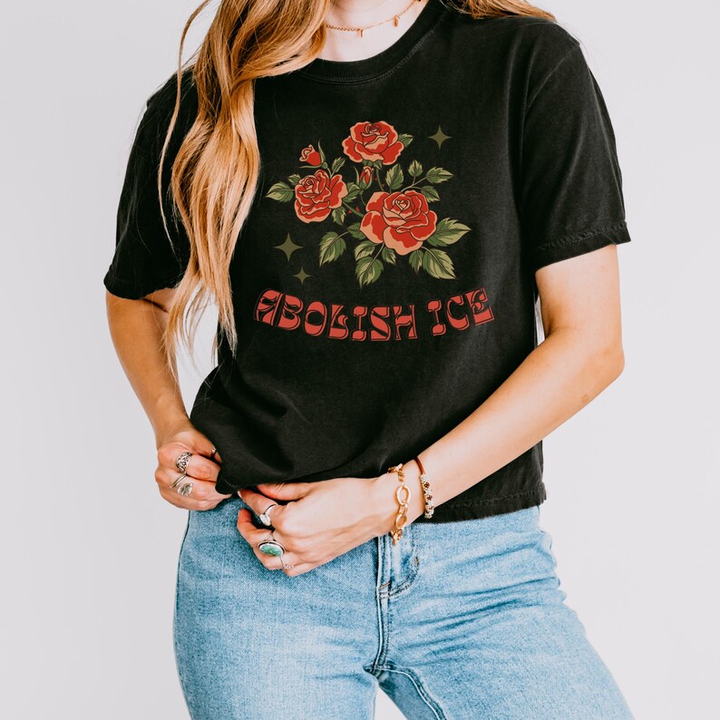 Woman in black cropped tee with red rose bouquet and “Abolish ICE” in retro font, styled with jeans and rings