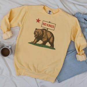 Yellow Comfort Colors sweatshirt flatlay with NO KINGS California bear graphic, paired with denim.