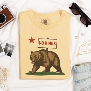 Yellow folded Comfort Colors t-shirt with bear holding NO KINGS sign, styled with camera and sunglasses.