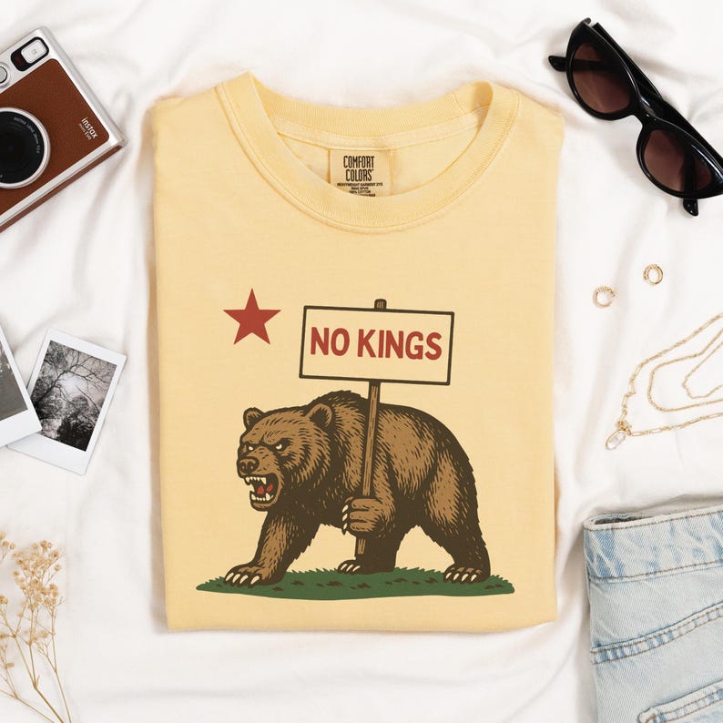 Yellow folded Comfort Colors t-shirt with bear holding NO KINGS sign, styled with camera and sunglasses.