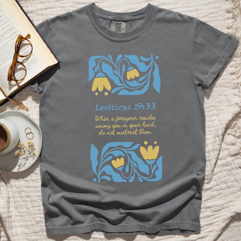 Pepper-colored tee with floral Leviticus 19:33 scripture graphic and immigrant advocacy message, shown on cozy bedspread