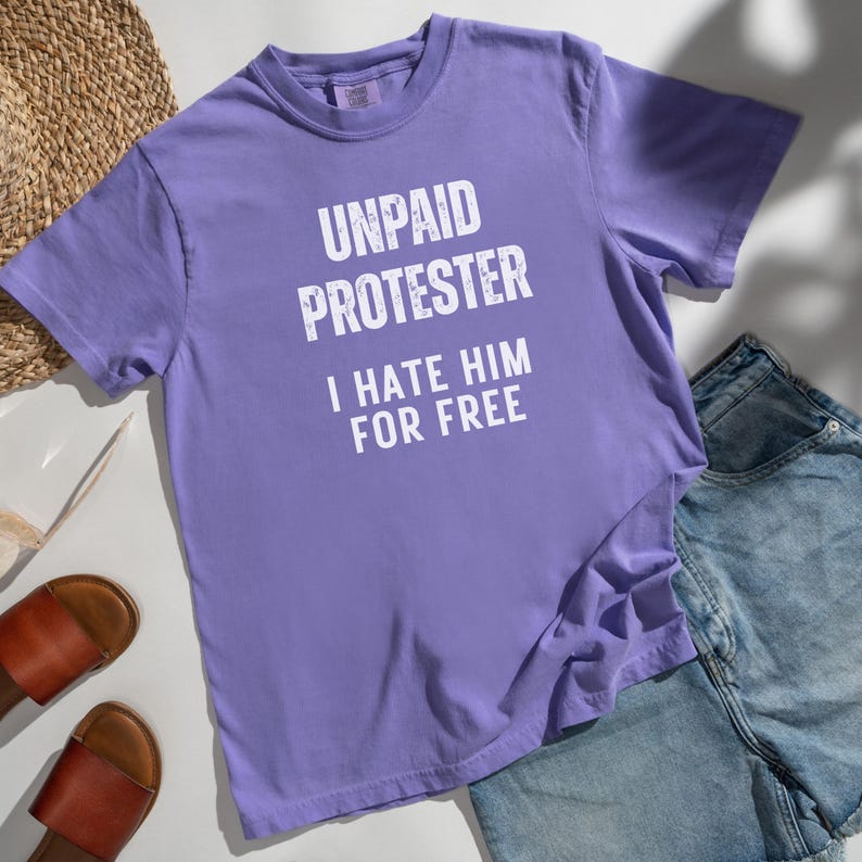 Lavender Comfort Colors tee with bold anti-Trump protest message laid on white blanket with straw hat and sandals.