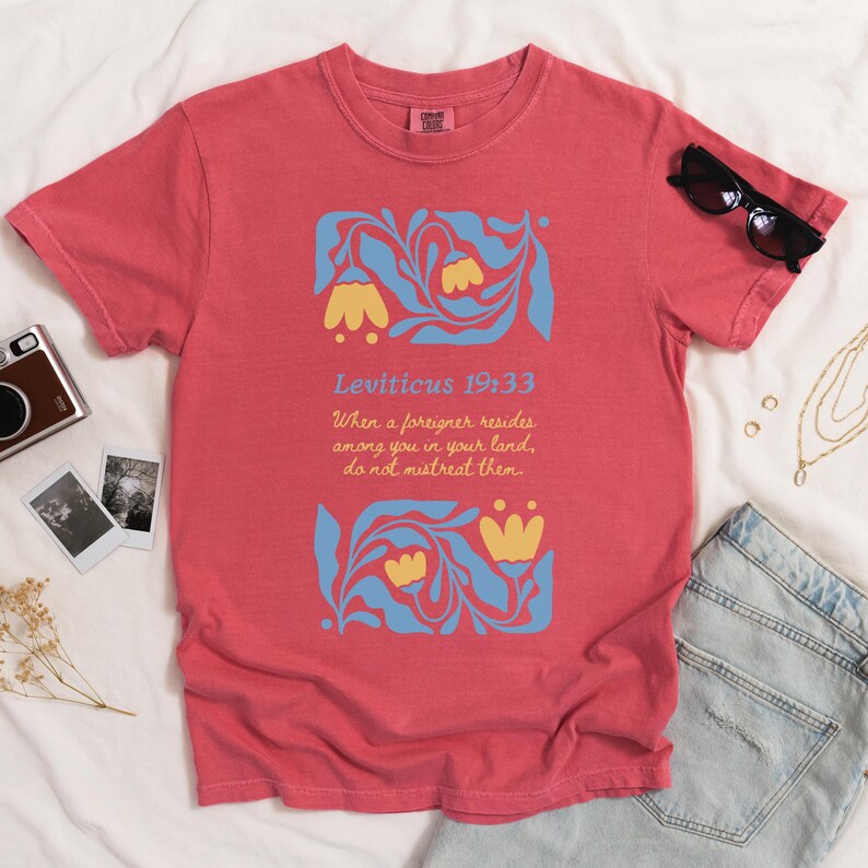 Watermelon pink comfort colors T-shirt laid flat with Leviticus 19:33 Bible quote in blue and yellow