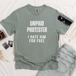 Flat lay of sage green Comfort Colors tee with the same text design, surrounded by denim and sunglasses.