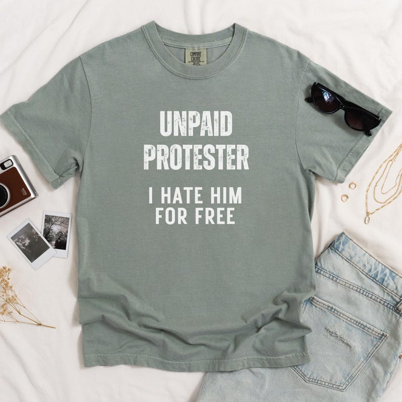 Flat lay of sage green Comfort Colors tee with the same text design, surrounded by denim and sunglasses.