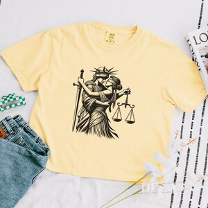 Pale yellow cropped tee flat lay with LGBTQ+ themed line drawing of two iconic female figures in romantic embrace
