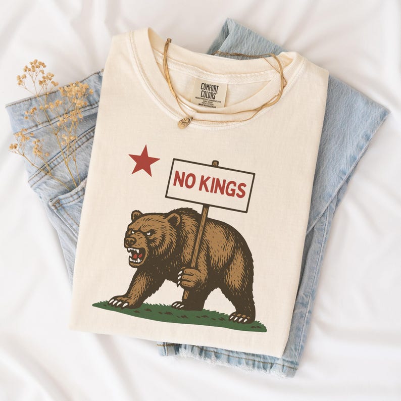 Natural folded Comfort Colors t-shirt with NO KINGS bear protest graphic, styled with denim and necklace.
