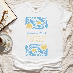 White Comfort Colors tee featuring folk floral graphic with Leviticus 19:33 Bible verse about immigrants, photographed on a neutral bedspread