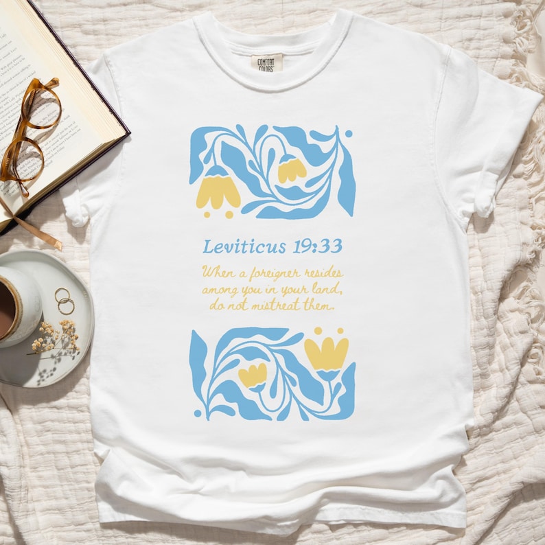 White Comfort Colors tee featuring folk floral graphic with Leviticus 19:33 Bible verse about immigrants, photographed on a neutral bedspread