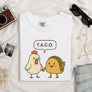 Puede incluir: Camiseta blanca con un pollo de dibujos animados y un taco tomados de la mano. El taco hace la señal de la paz. Encima de los personajes hay un bocadillo con las letras "T.A.C.O." impresas en negro. La camiseta es de material suave.