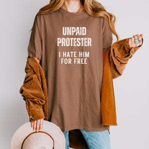 Person wearing brown Comfort Colors tee with bold white text that says “Unpaid Protester – I Hate Him For Free” in a modern anti-Trump protest graphic style.