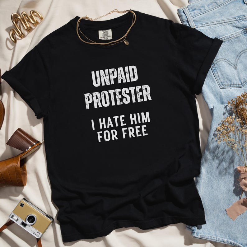 Comfort Colors tee with bold anti-Trump protest message laid on white blanket with straw hat and sandals.