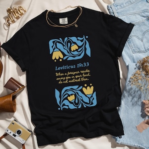 Black Comfort Colors T-shirt with blue and yellow floral art and Bible verse from Leviticus 19:33, styled with accessories for Etsy listing