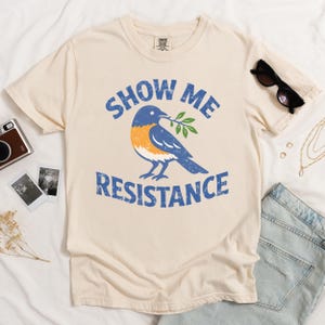 以下が含まれることがあります： 「SHOW ME RESISTANCE」の青い文字と、オリーブの枝を持った青い鳥のグラフィックが特徴のクリーム色のTシャツ。 サングラス、ジュエリー、デニムショーツとコーディネートされています。