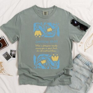 Bay green Comfort Colors T-shirt with retro Bible verse design reading “Do not mistreat them” and Matisse-style florals in yellow and blue